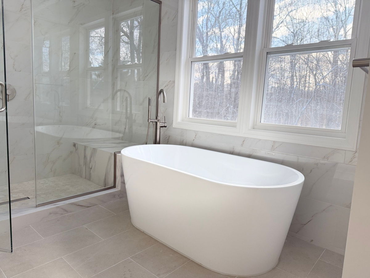 Warren master bathroom remodel featuring a freestanding white soaking tub, marble-look tile surround and flooring, frameless glass shower enclosure, and large windows overlooking wooded landscape.