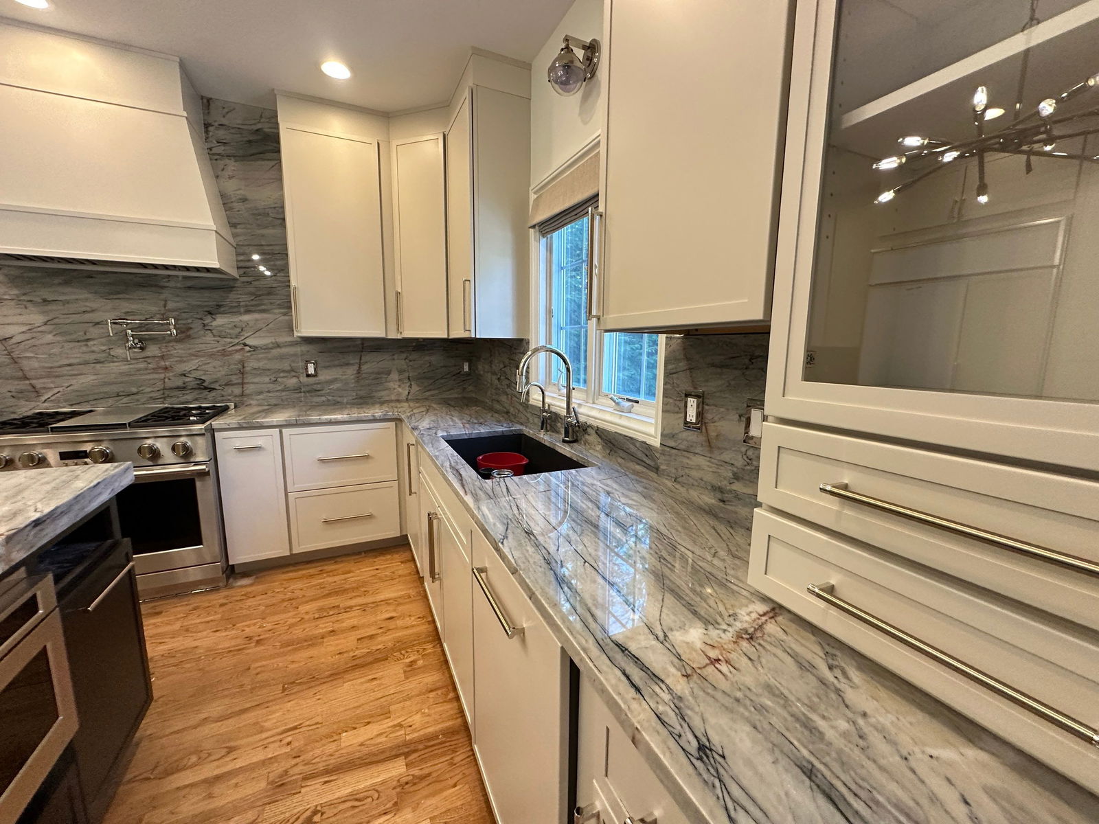 Galley kitchen with cream shaker cabinets, gray marble veined countertops and backsplash, stainless steel appliances, undermount sink with chrome faucet, wood plank flooring, and recessed ceiling lights.