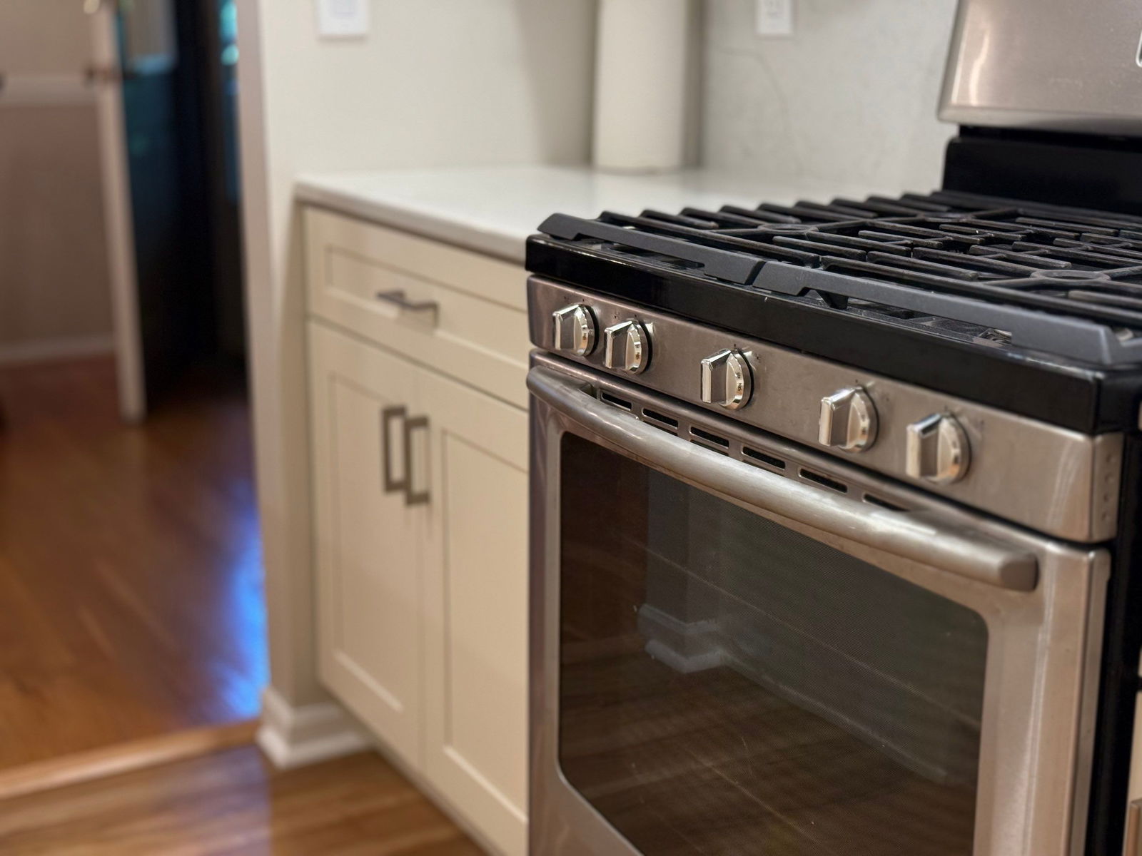 Stainless steel gas range with black cooktop and five knob controls beside cream-colored shaker cabinetry in a kitchen with hardwood flooring.